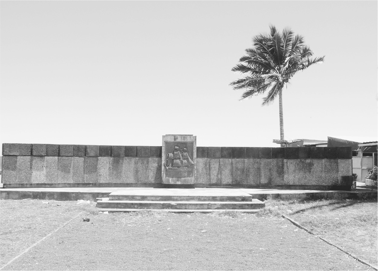 A photograph of Monument to the 1665 settlement of Réunion. It has a tall tree after the wall and a statue on the wall.