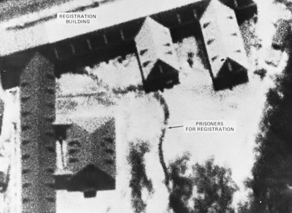 A hazy aerial image of a concentration camp featuring labeled buildings and a line of prisoners for registration.