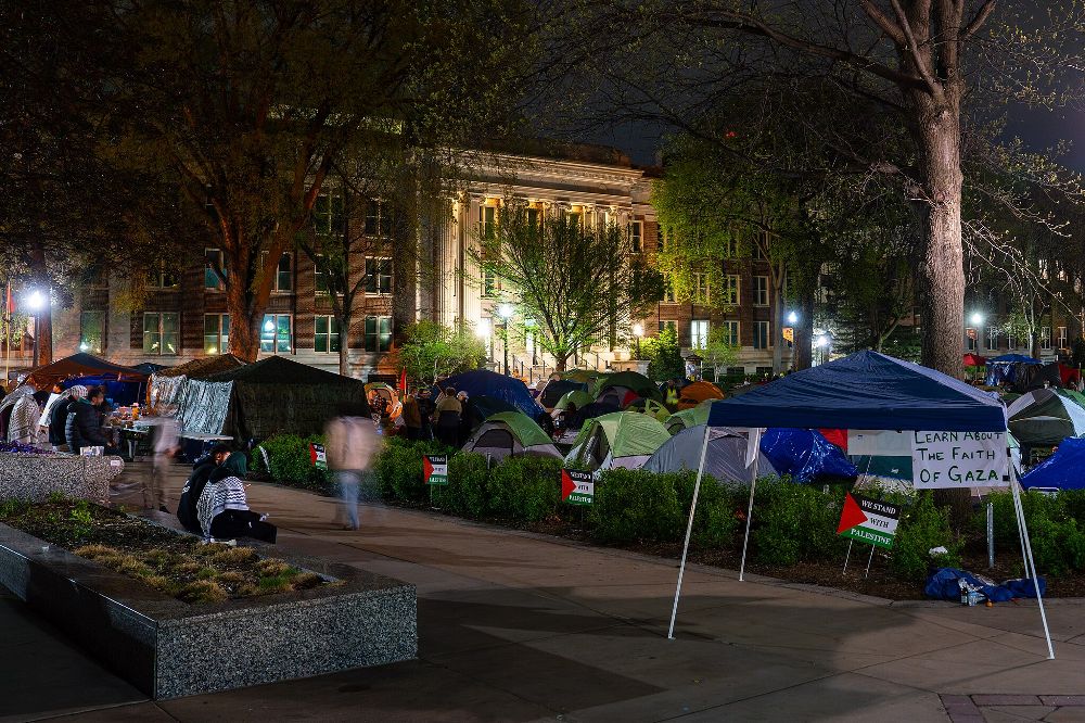 Tents pitched on the lawn of the University of Minnesota at night. Lawn signs that read “We stand with Palestine” surround the tents. In the background are campus buildings with Greek columns.