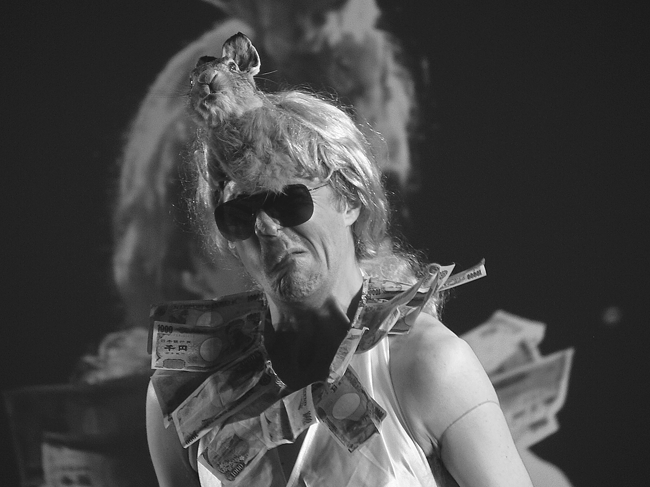 Marcus Coates wearing sunglasses, a wig, and a costume adorned with paper money, with a small animal perched on their head.