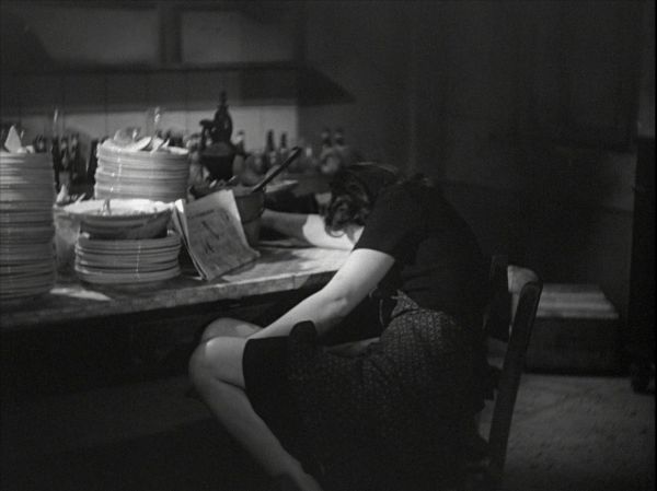 Woman seated at kitchen table, resting her head on her arm. Stacks of dishes and a newspaper in front of her on table.