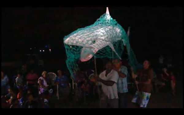 Nightime image of a papier-mâché shark covered in netting being paraded about.