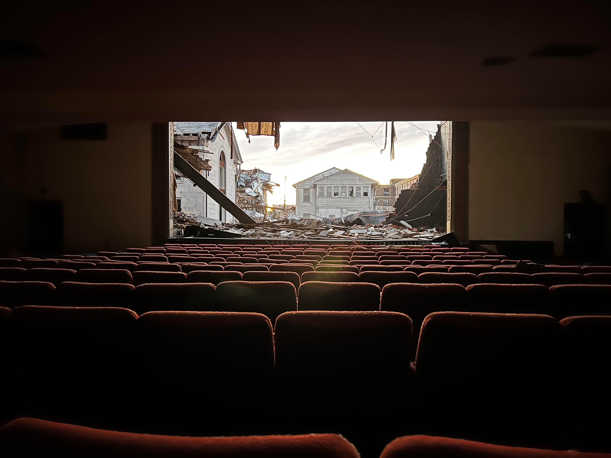 View from inside a movie theater showing rows of seats and a view to rubble and destroyed buildings outside through a missing wall where the theater screen would normally be.