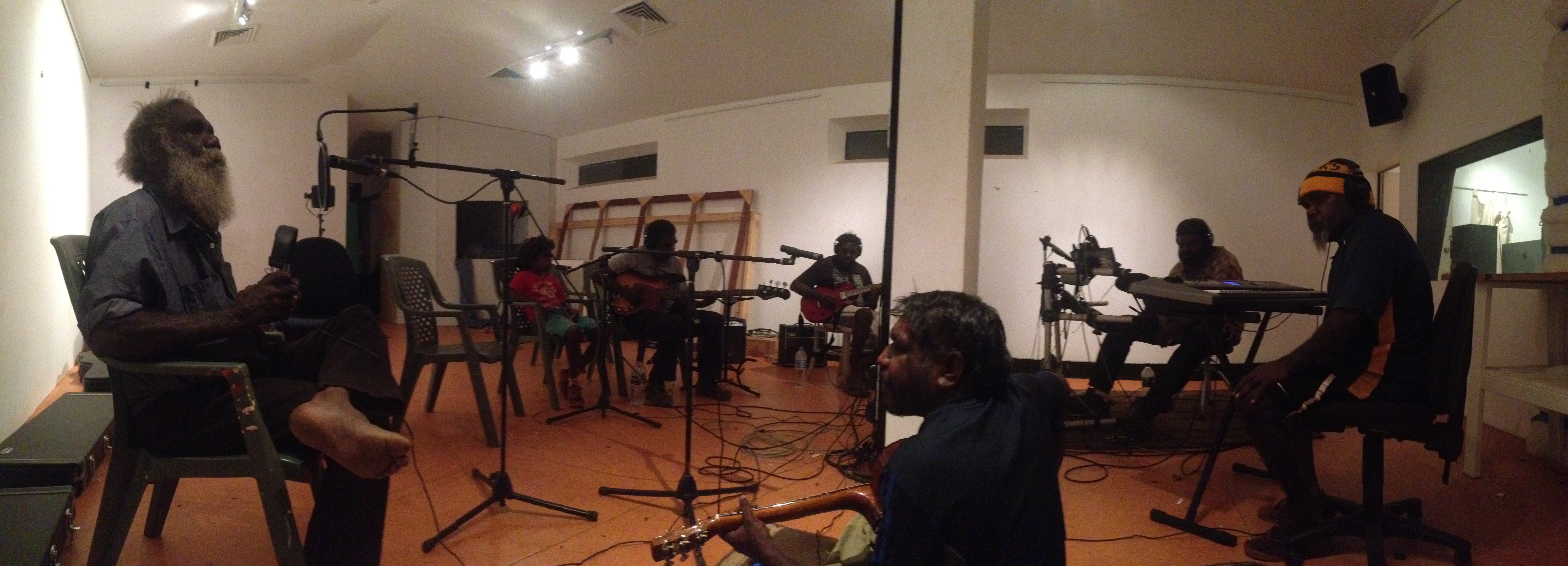 Panoramic view of a music studio with seven people sitting in a large circle with instruments; at left, Mark is singing into a microphone.