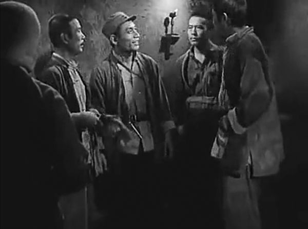 Black-and-white still of a group of five men talking amongst themselves in close quarters.