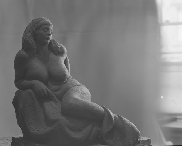 Frontal view of a clay sculpture of a voluptuous, reclining nude female figure whose feet are covered by drapery.