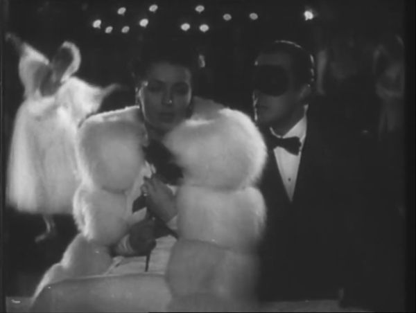 A woman, elegantly dressed in a white fur coat, walks with a man wearing a tuxedo and mask as an outdoor nighttime ballet takes place behind them.