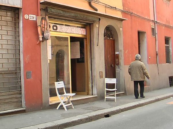 A plain storefront, with an open door and two folding chairs on the sidewalk beside it; the only person depicted, a middle-aged man, is looking away from the doorway.