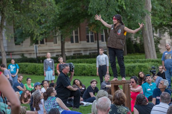 A woman in a brown vest with red lines drawn across her eyes stands on a high stool, arms out, talking. People sit around her on the grass, watching.