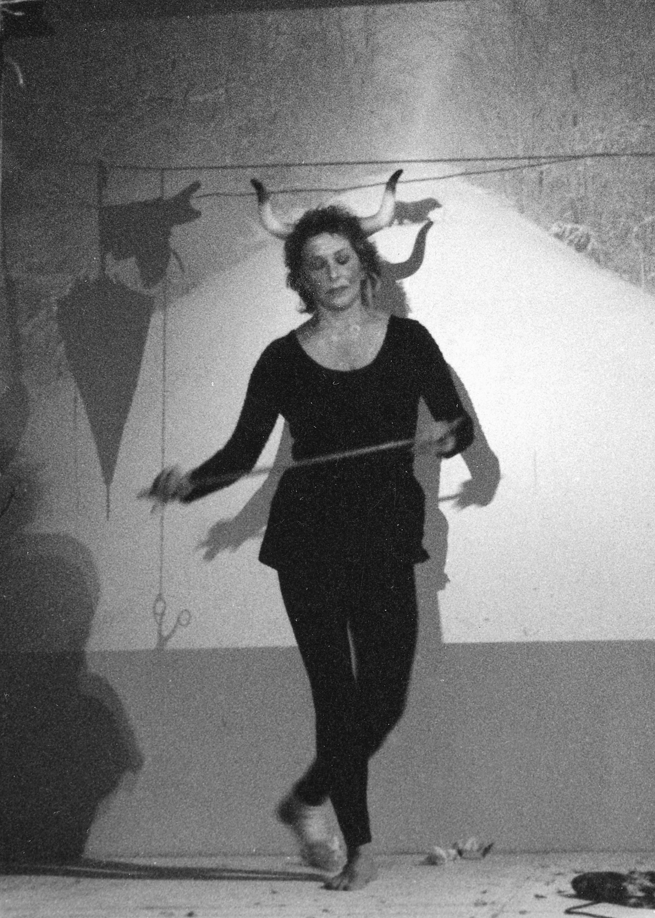 Carolee Schneemann wearing a headpiece with horns, performing with a stick in a dimly lit space.