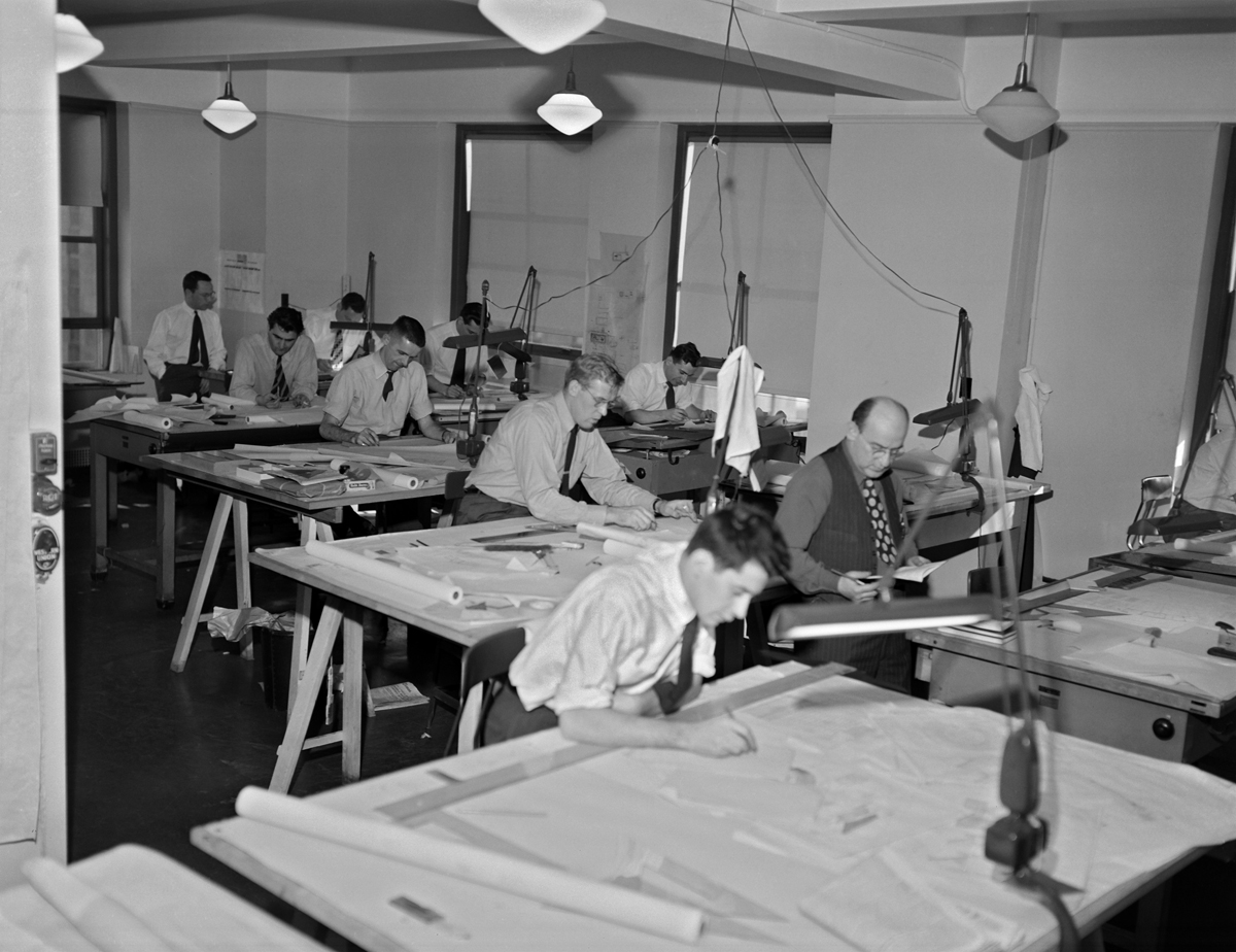 Photograph of drafting room with nine men in shirtsleeves and ties working on drawing boards.