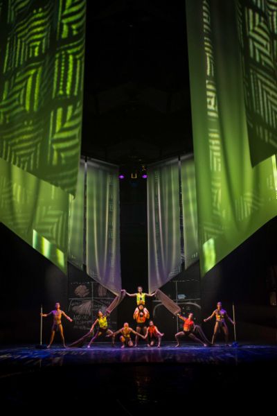 Long angled curtains hang down. Beneath eight dancers form a house. Two on the ends hold mops, two are stacked in center, four stretch arms out.