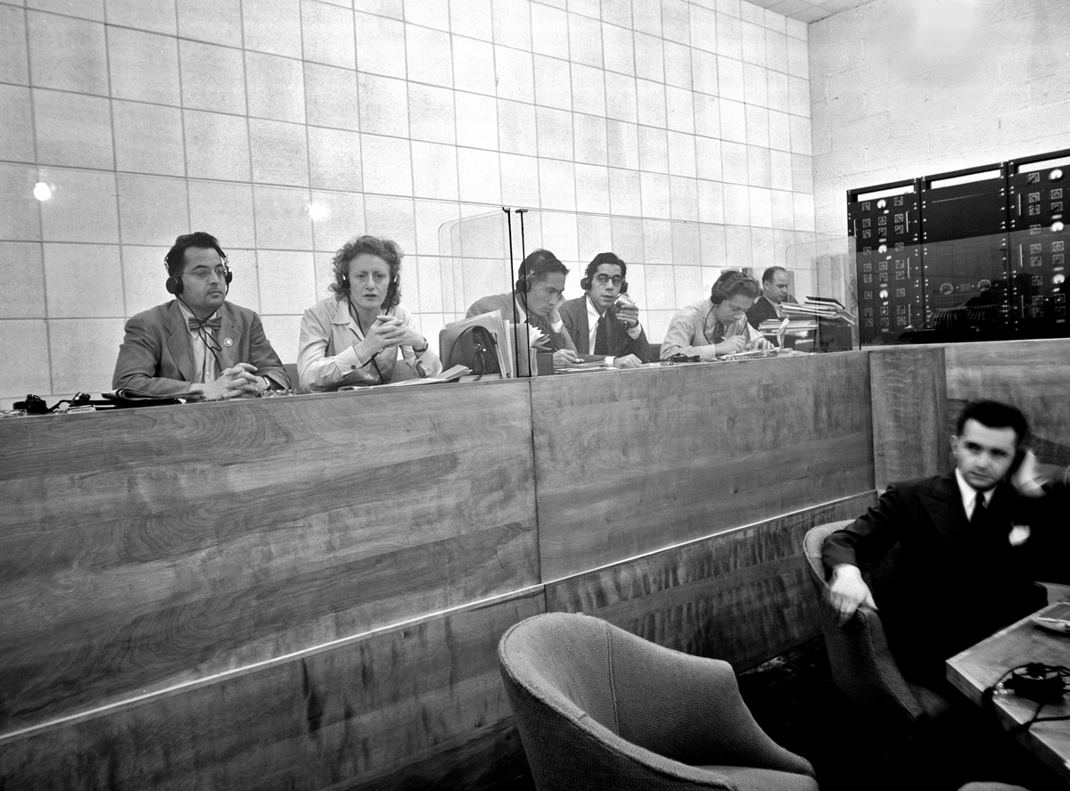 Photograph of male and female interpreters wearing headphones behind glass panels and in front of a tiled wall.