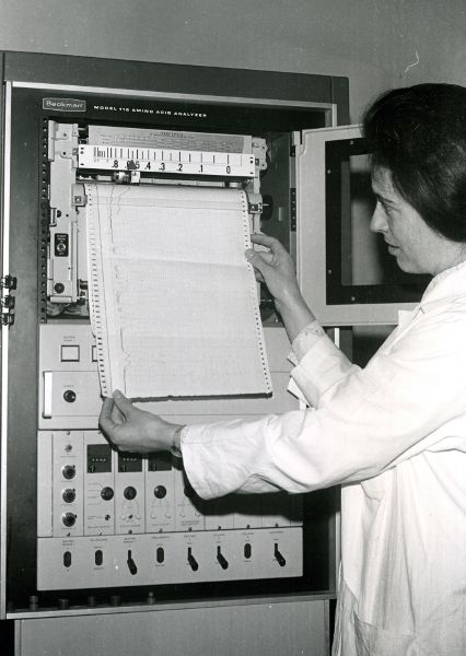 A technician wearing a white lab coat holding a paper readout that is being printed from person-sized piece of equipment.