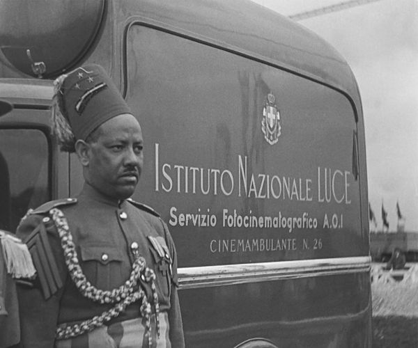 A Black man in a military uniform stands stiffly next to a LUCE Institute mobile projection van.