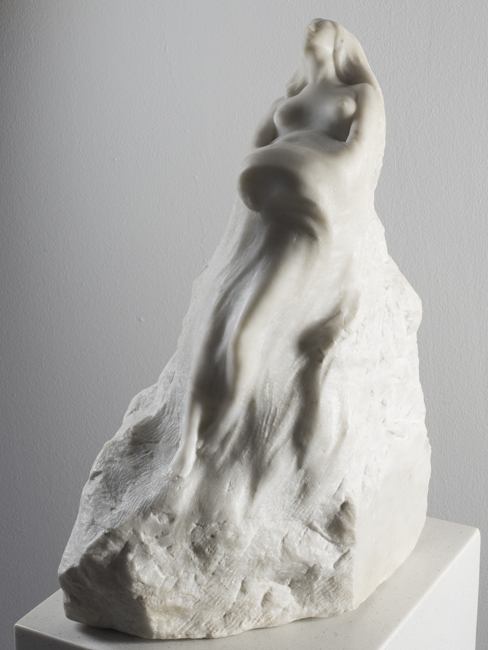 The exhibit features Franz Flaum’s “Wave (Morning).” This marble sculpture, with a flat base and a non-regular shape, points upwards, suggesting a woman lying on her back. The intricately carved piece showcases detailed craftsmanship on marble.