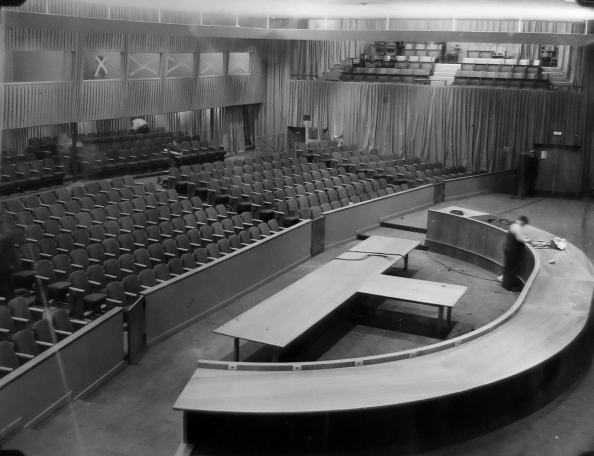 Photograph of an empty council chamber under construction with a man working on a large semicircular plywood table facing auditorium-style seats.