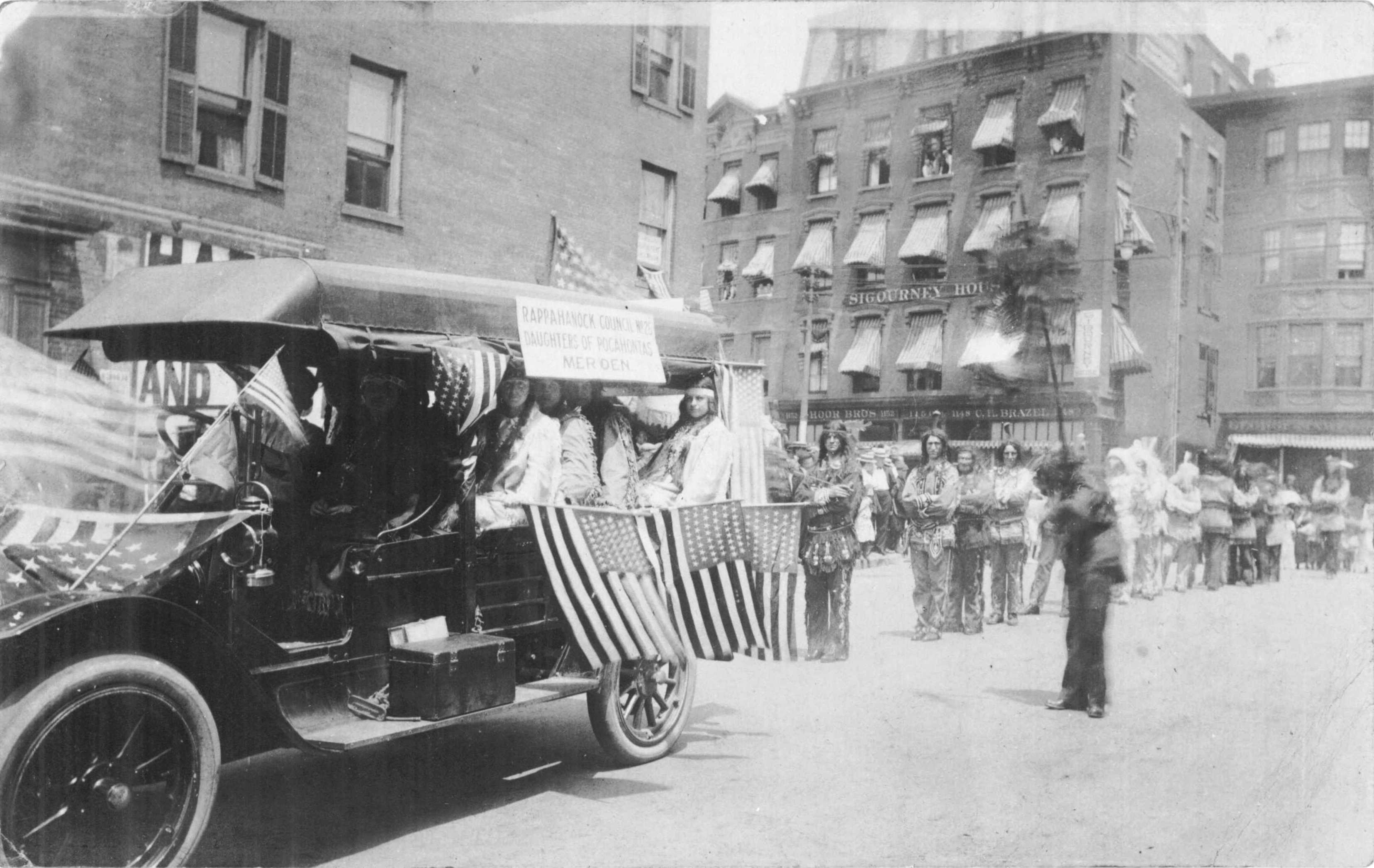 Several white women in Native American costumes sit in an old car adorned with American flags followed by a line of white men in similar costumes.