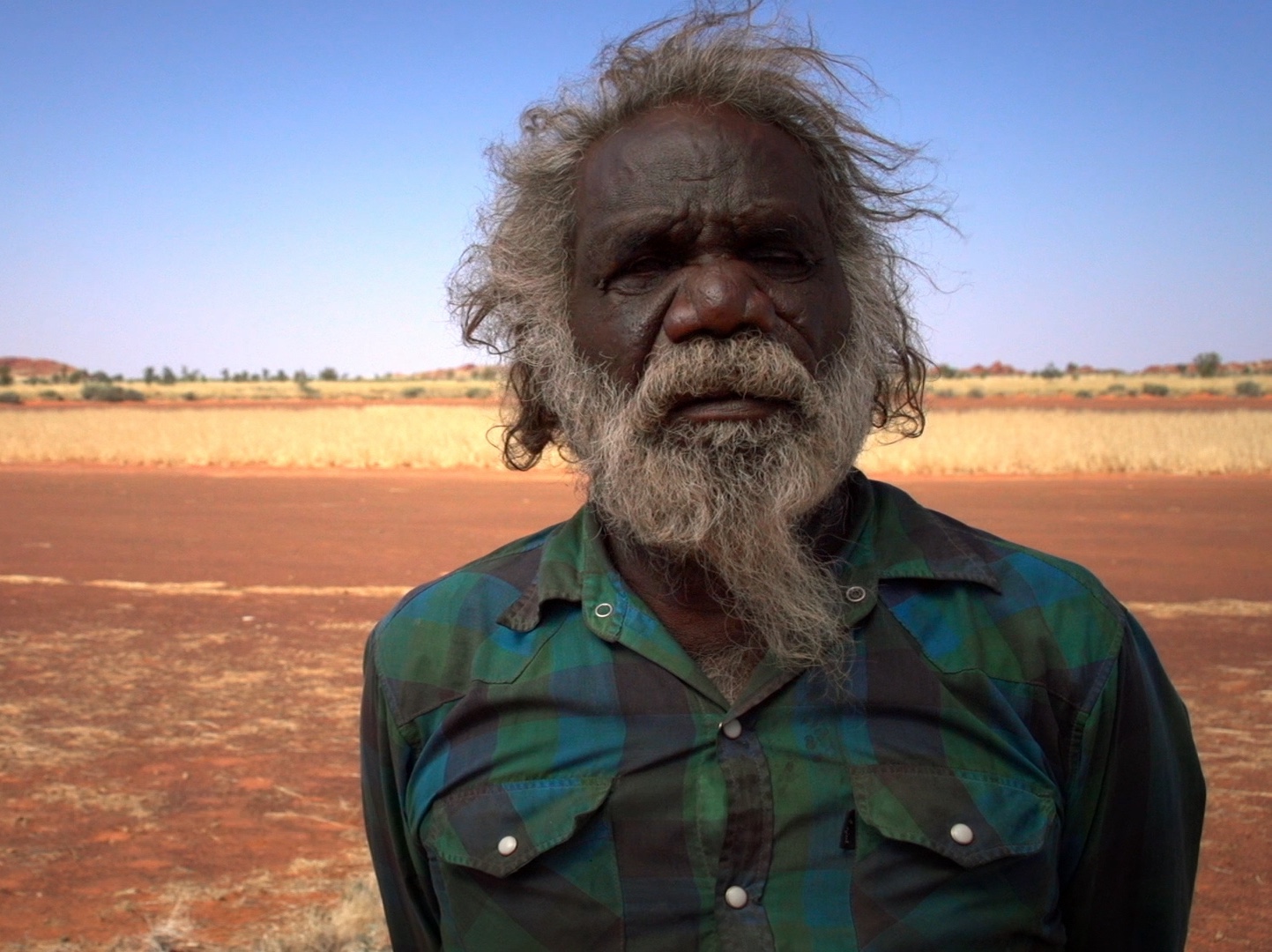 Mark’s upper body and face take up the right side. He looks directly into camera in front of an orange clay ground and blue sky.