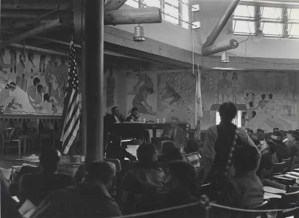 A man, seen from behind, stands in a room decorated with murals, with seated people facing two men sitting on a raised dais.