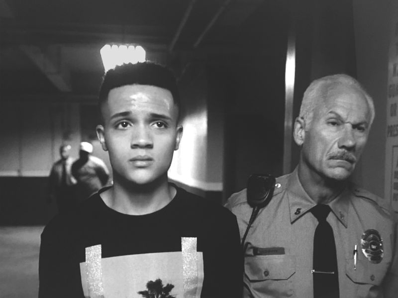 A young person with a concerned look on his face walks beside a middle-aged police officer in a poorly lit concrete structure.