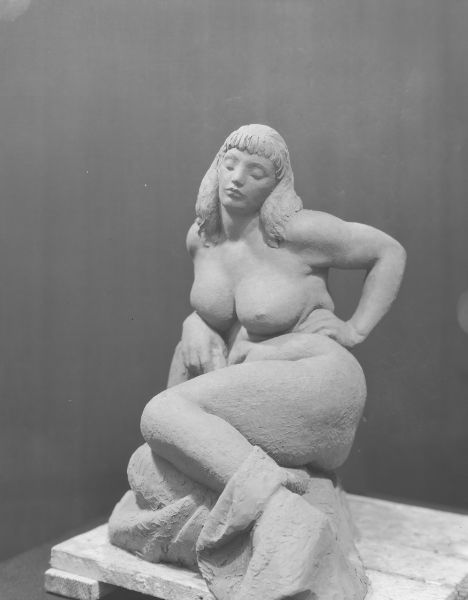 Foreshortened view from the feet of a clay sculpture of a voluptuous, reclining nude female figure.