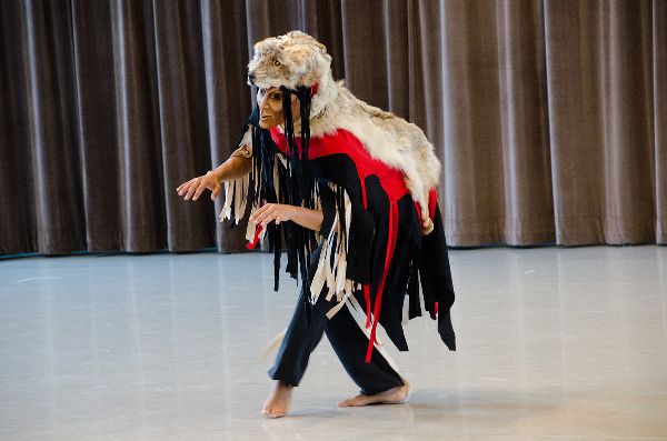A masked dancer, wearing a wolf pelt on their head and shoulders, stands center stage, arms forward with fingers down, legs crossed, crouching.