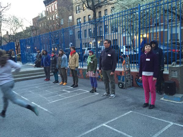 On an asphalt playground, people dressed warmly stand before a fence, evenly spaced, wearing name tags. A blurry figure runs out of the frame.