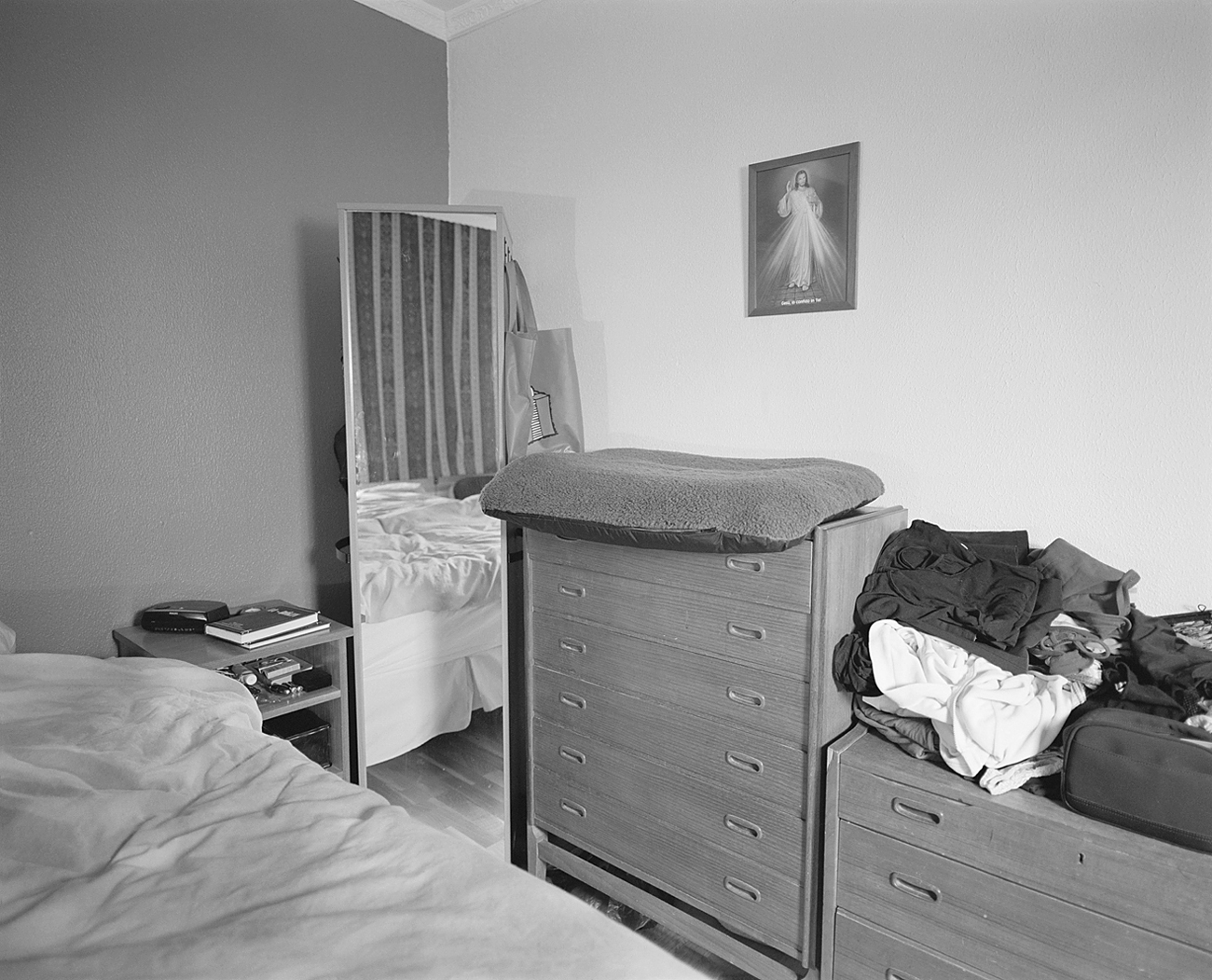 A bedroom with a dresser, a mirror reflecting a bed, and clothes stacked on furniture.