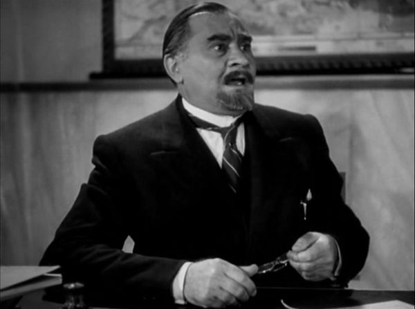 A male professor in a suit sits at his desk. Holding his glasses in his hands, he, in shock and disbelief, watches a scene unfold to his left.