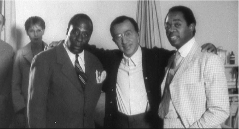 The exhibit showcases three men dressed in formal attire, posing together in a room. The man in the middle is seen placing his hands on the shoulders of the two on either side of him.