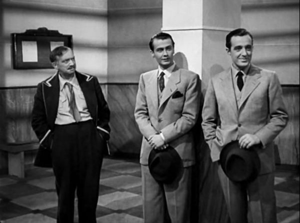 In a school hallway, two young men in suits stare at a figure outside the frame. Standing next to them is a janitor in uniform who looks at the men.