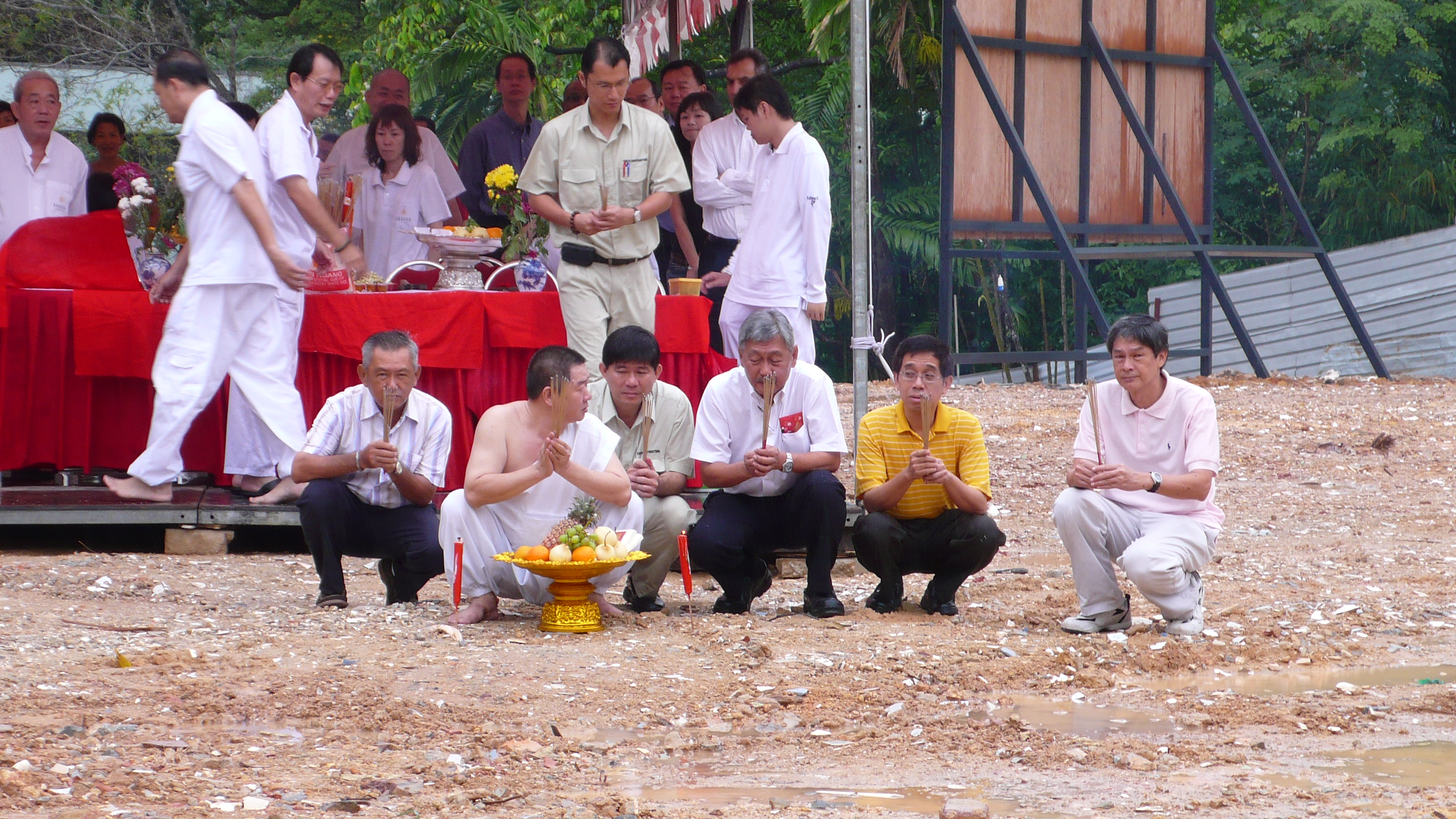 Several men kneel before a tent on the muddy ground of a construction site, holding joss sticks. At the center is a spirit medium in a white robe.