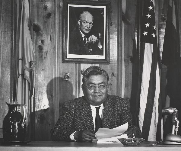A man with glasses at a desk beneath a portrait of the president of the United States, between two flags.