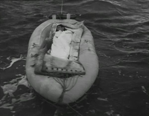 A man, wrapped in a white shroud, lies in the center of a small rescue boat as a larger vessel (not pictured) pulls the boat across the ocean.