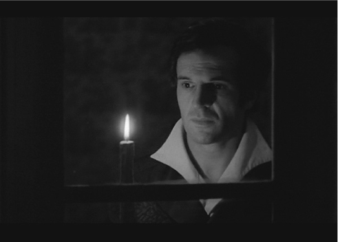 The exhibit portrays a man in a dim setting, his gaze fixed on a lit candle before him, and his expression communicates seriousness.