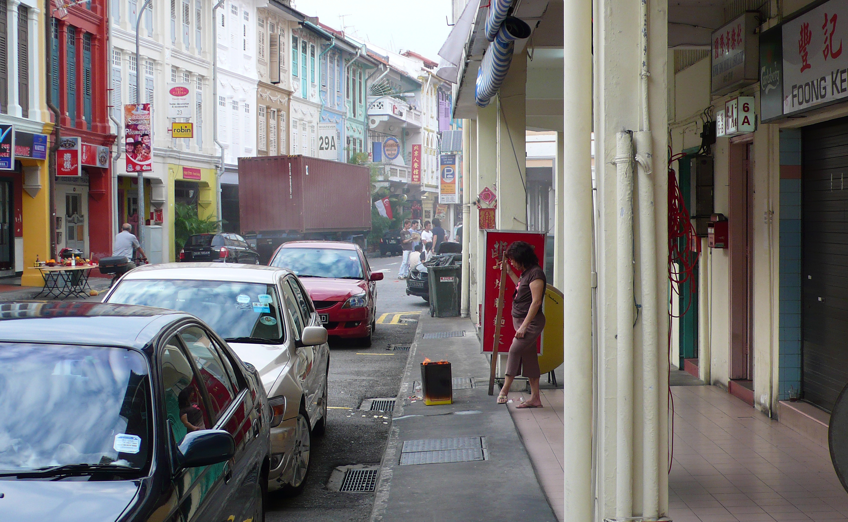 	A woman stands in front of a fire in a small metal can, on a smoky street in Singapore. Other ritual offerings are happening in the background.