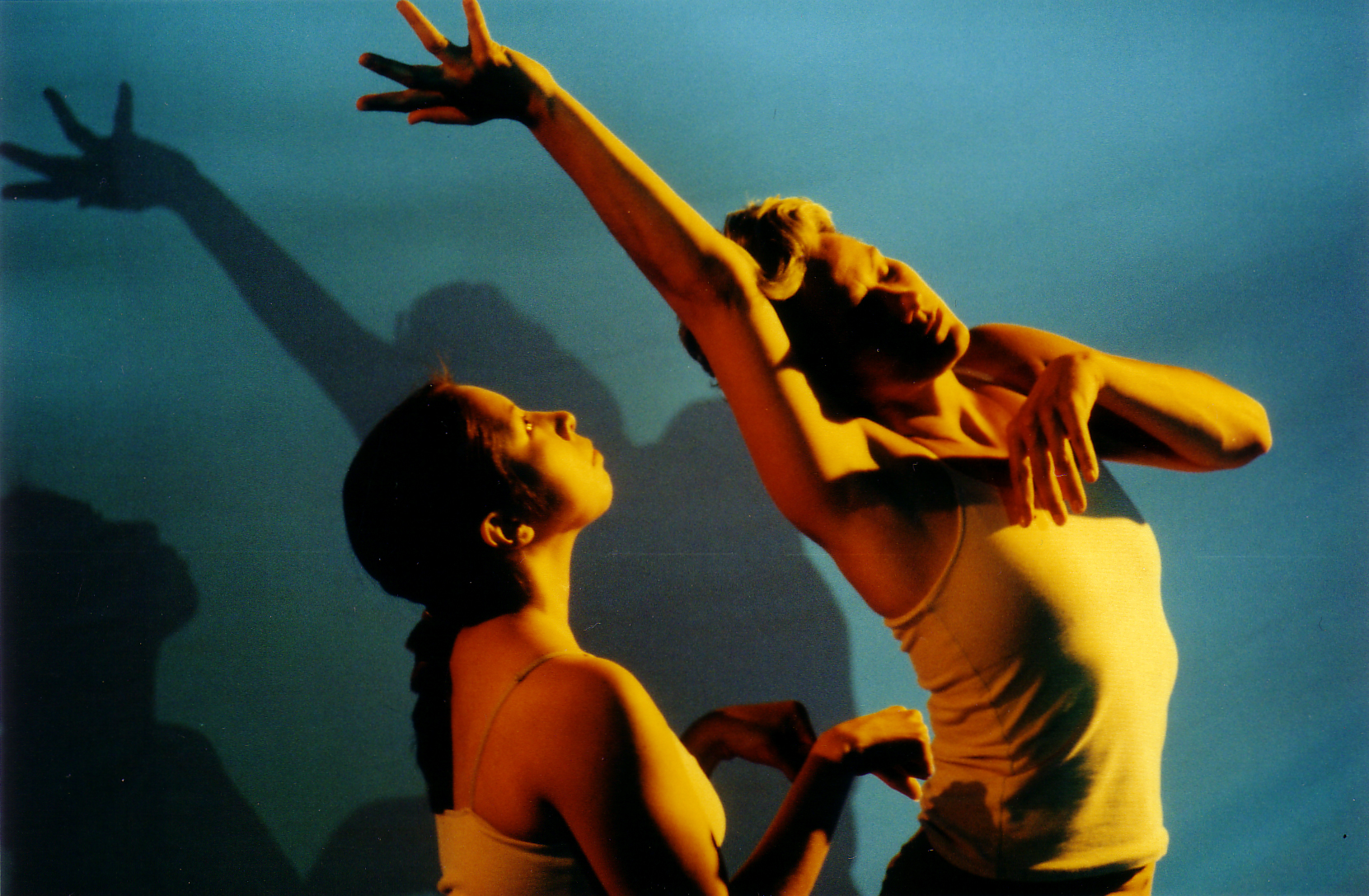 Two performers on stage beside one another, one extending their body upward with one arm raised and head lilting into it; the other performer gazes upward, hands, sligltly fisted and slightly extended at chest level.