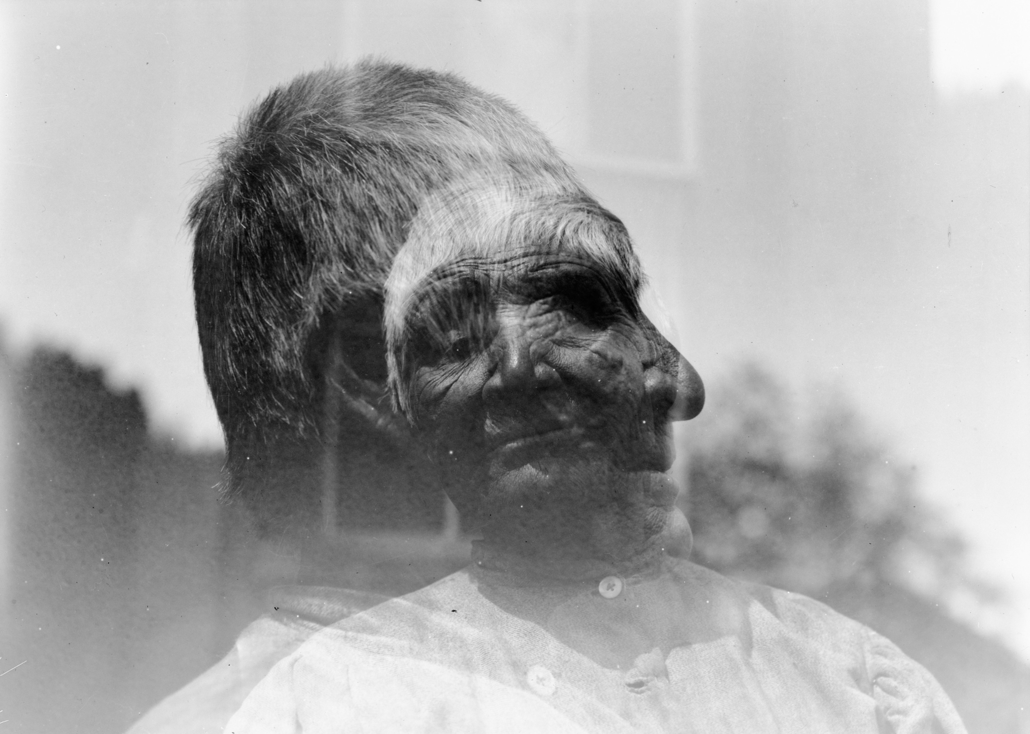Double-exposed image of facing and profile headshots of an older Native American woman. A third exposure faintly indicates a window frame.