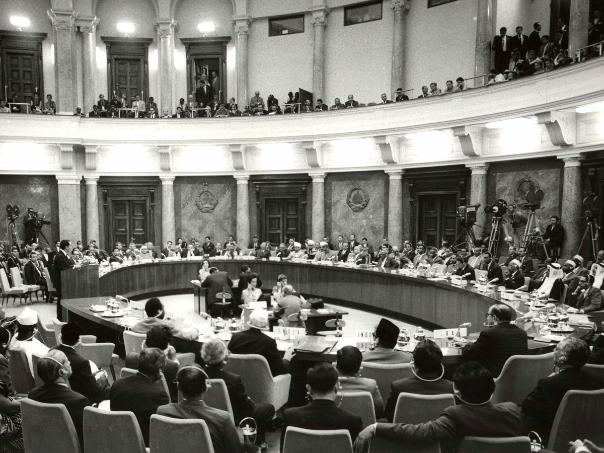 Photograph of convening delegates with headphones seated around an oval table in a two-tier room with columns.