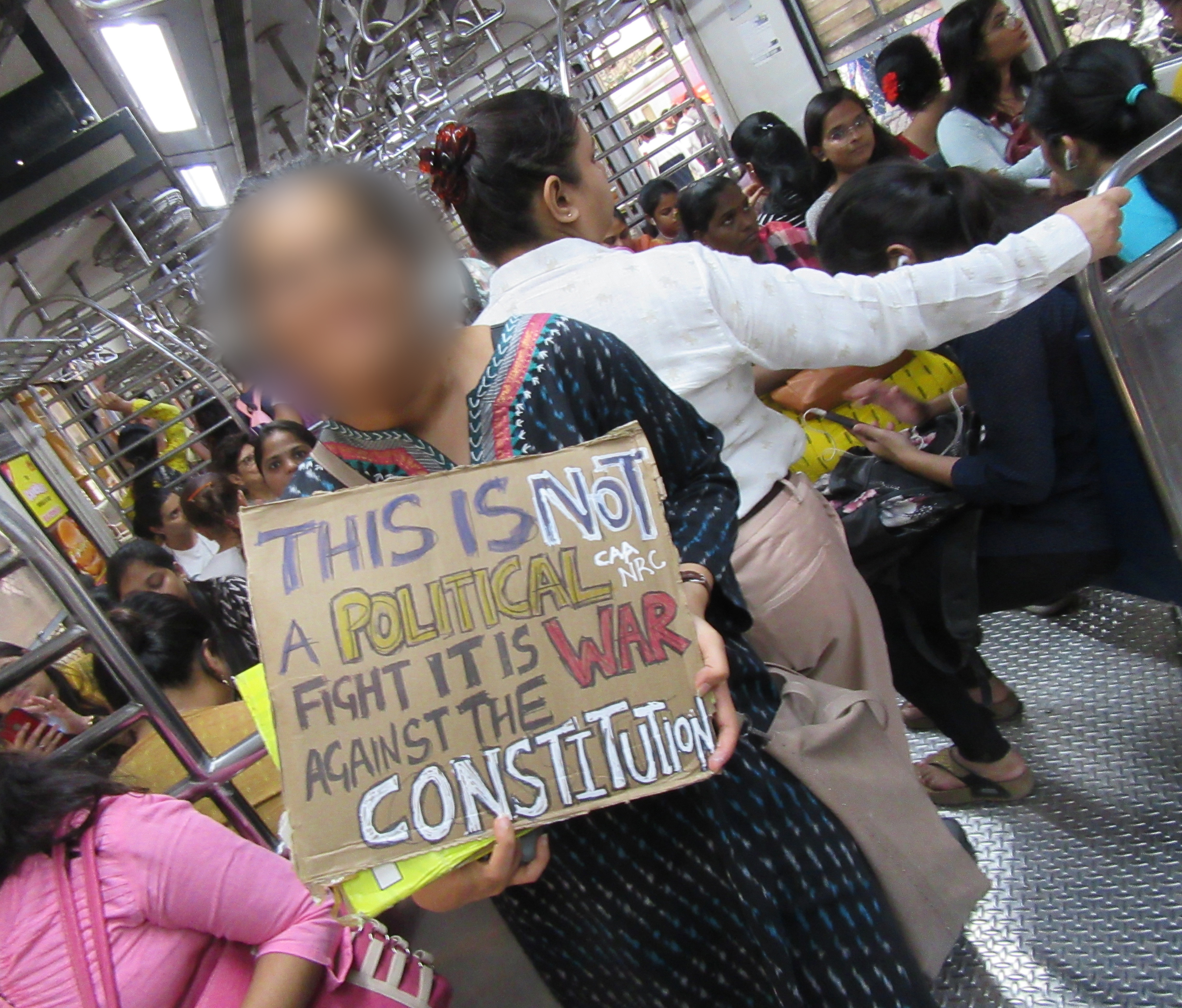 A woman standing on a public train holds a protest sign. The train is full of riders, standing and sitting.