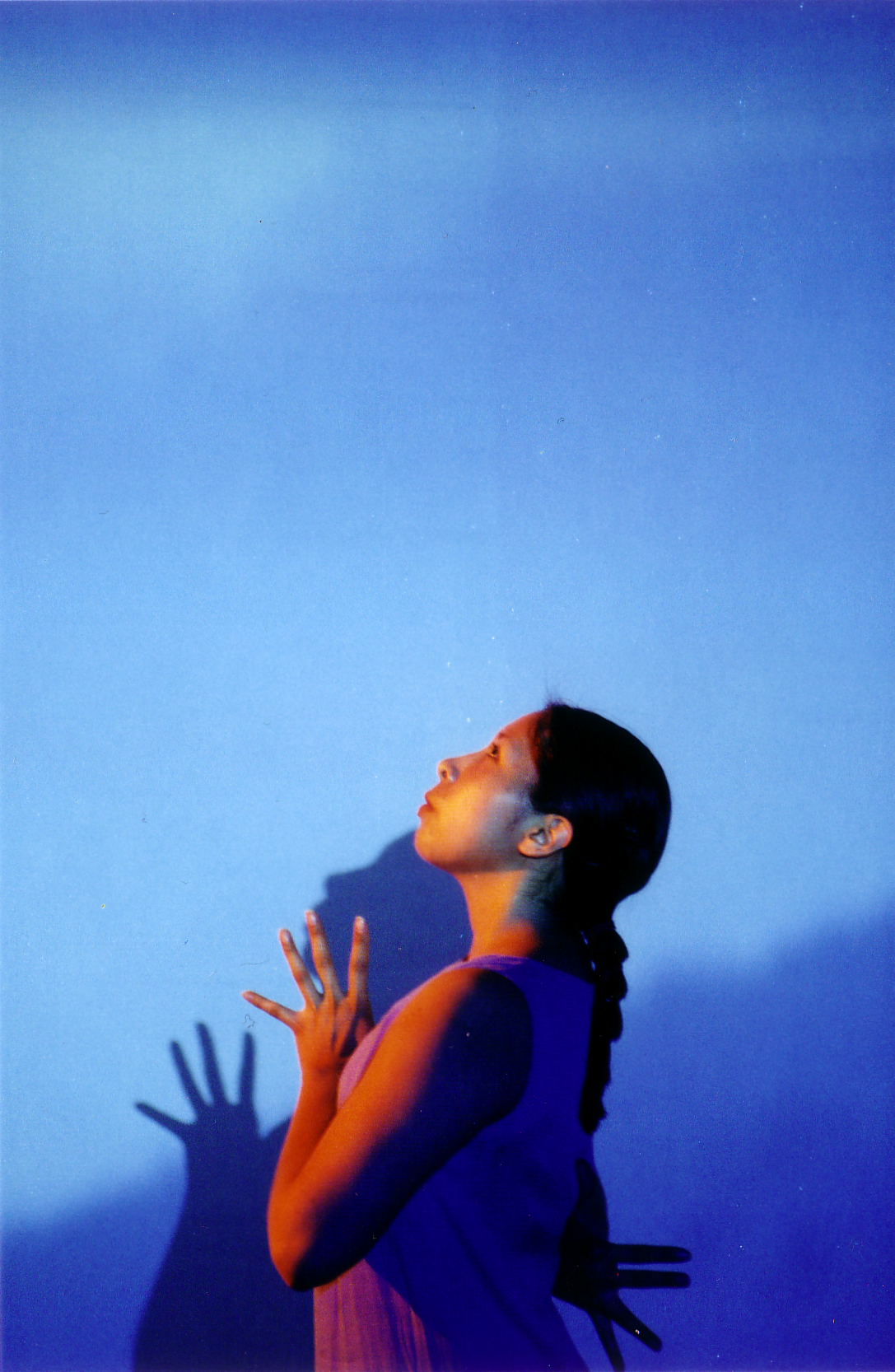Sidevide of a performer onstage in front of a blue backgrop, hands togeter at chest with fingers splayed and extended upward, following the performs’s gaze.
