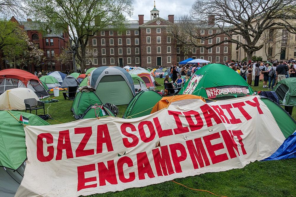 A large white banner reading “Gaza Solidarity Encampment” in large red lettering is pinned to tents pitched on the lawns of Brown University. A large group of students is gathered on the lawn between tents.
