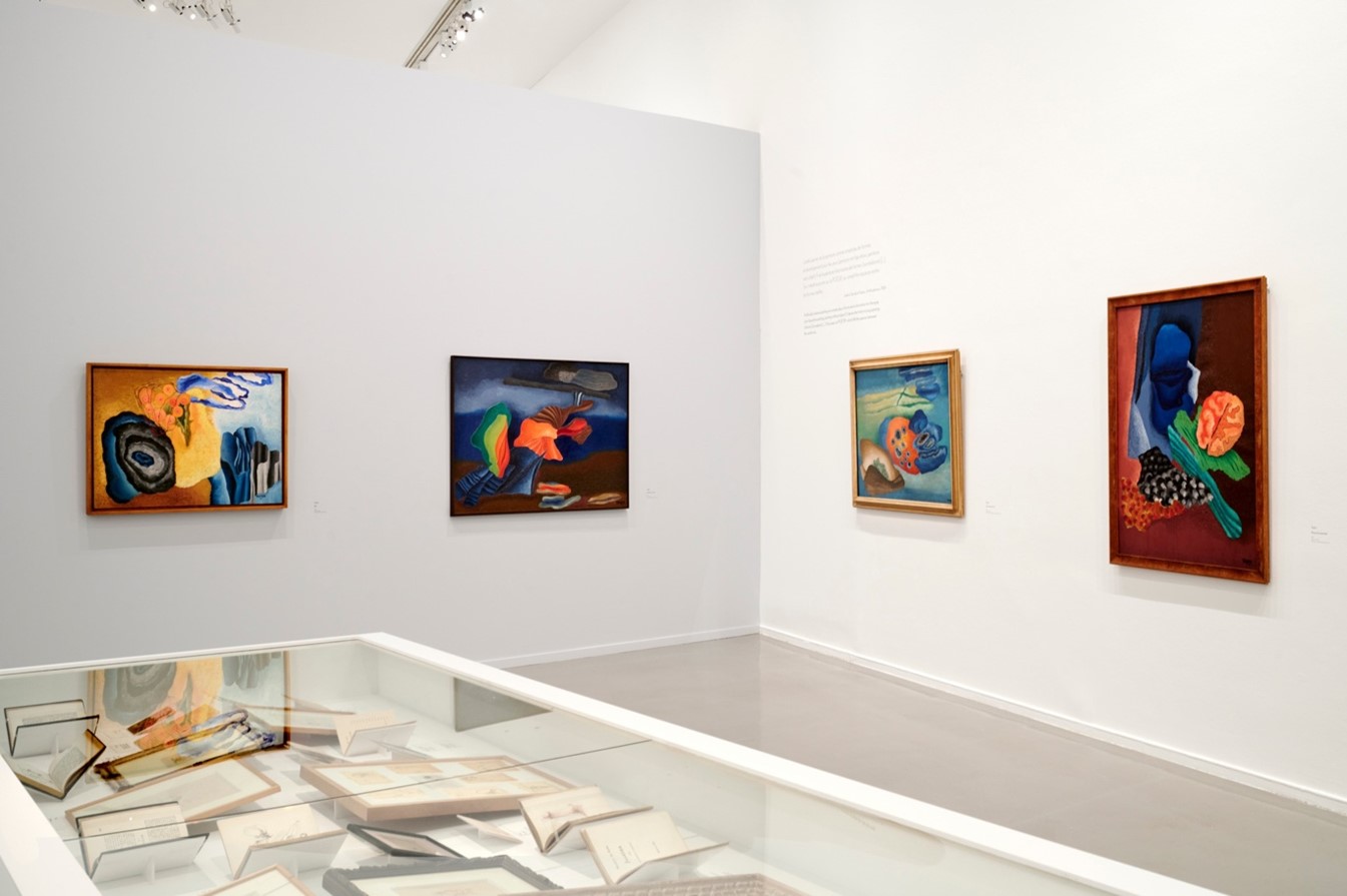 A glass case of documents. Behind it, four framed paintings on a white wall.