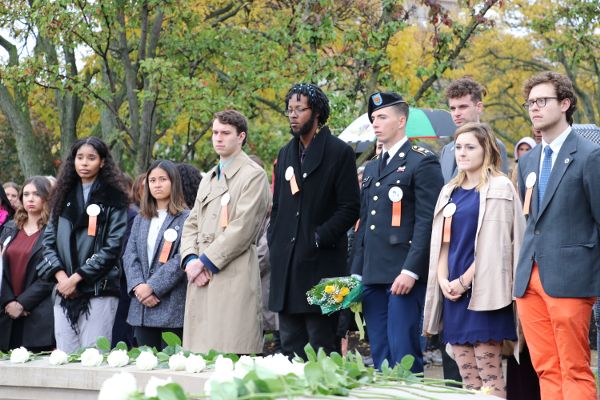 College-aged people with somber expressions stand side by side. Single white roses are lined up on a low wall in front of them.