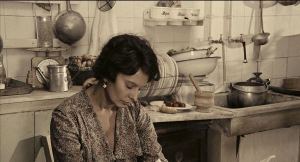 Woman sits at kitchen table with closed eyes, presumably asleep. Kitchen paraphernalia behind her on counter and in sink.