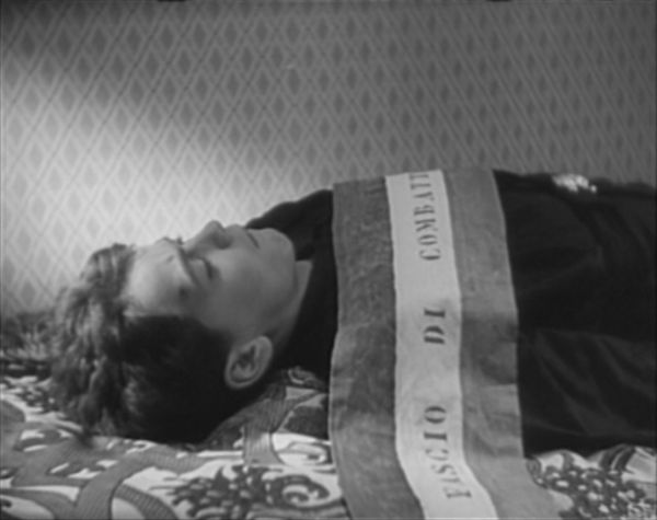 The body of a young man rests in a casket. He wears black and bears a banner across his body that announces he is a member of the Fascist Party.