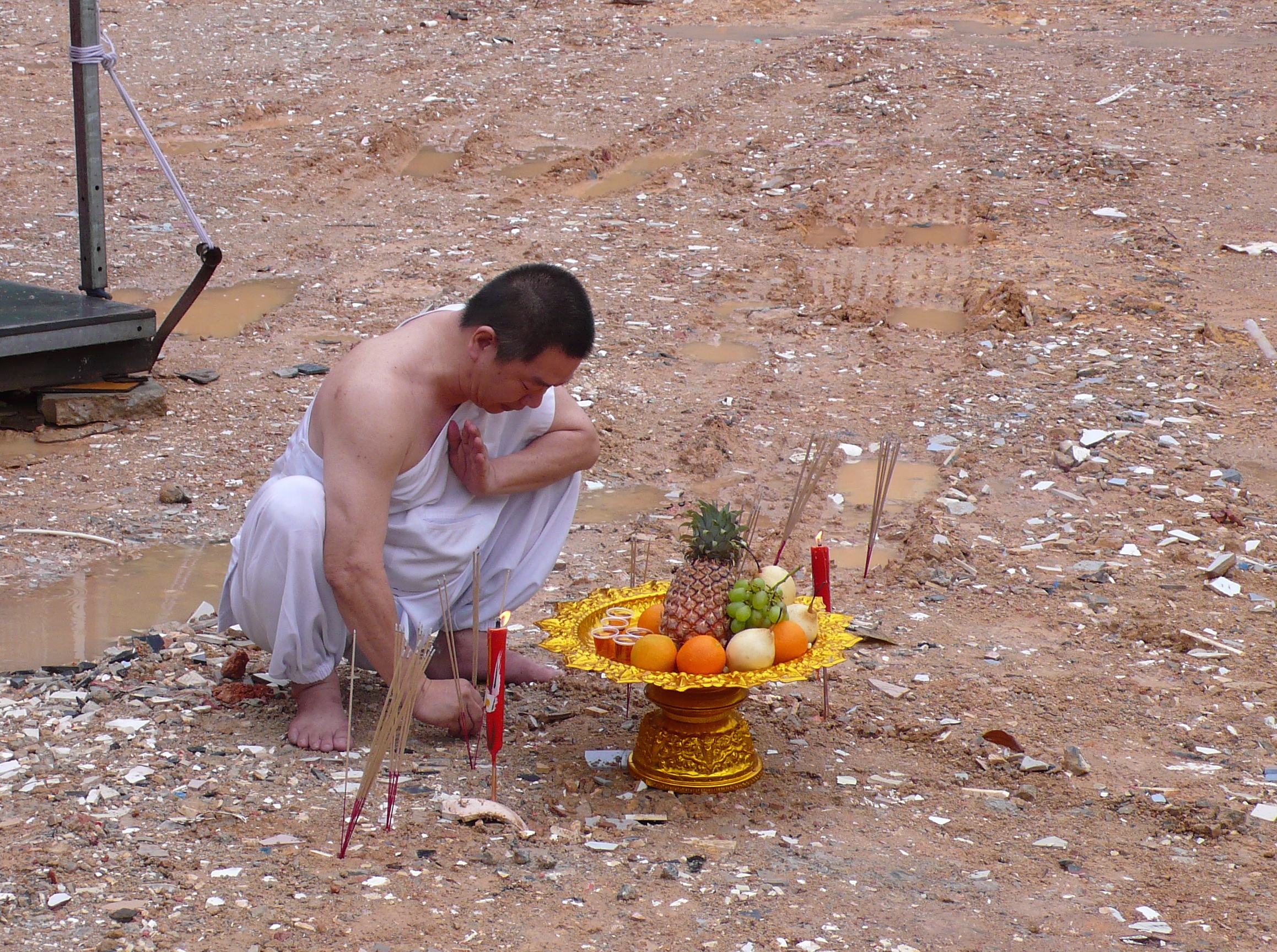 A Chinese man, a spirit medium, squats barefoot on the earth behind a gold plate. His head is bowed, and the side of one hand is at his chest.