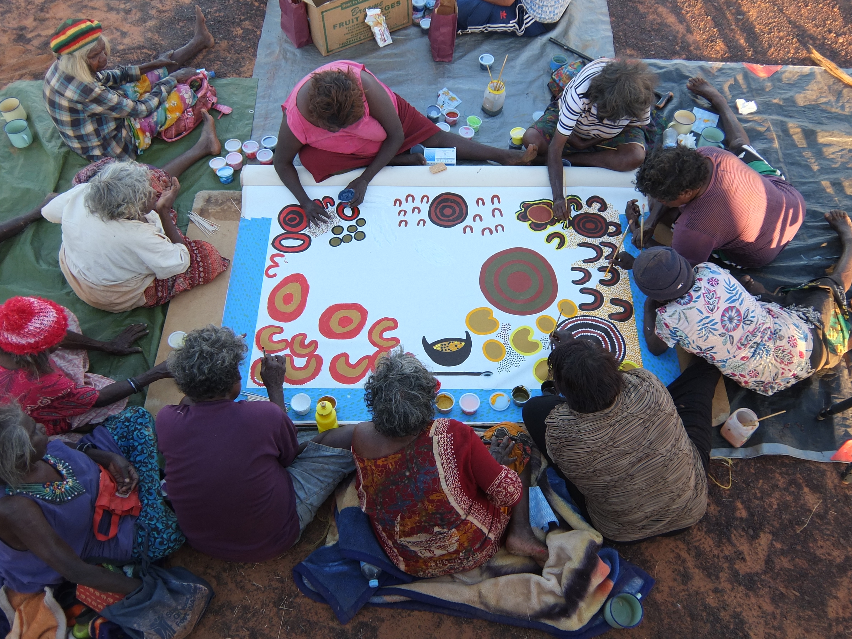 In a view from above, eight women are sitting around the edge of a large, half-finished canvas as each paints a different section.