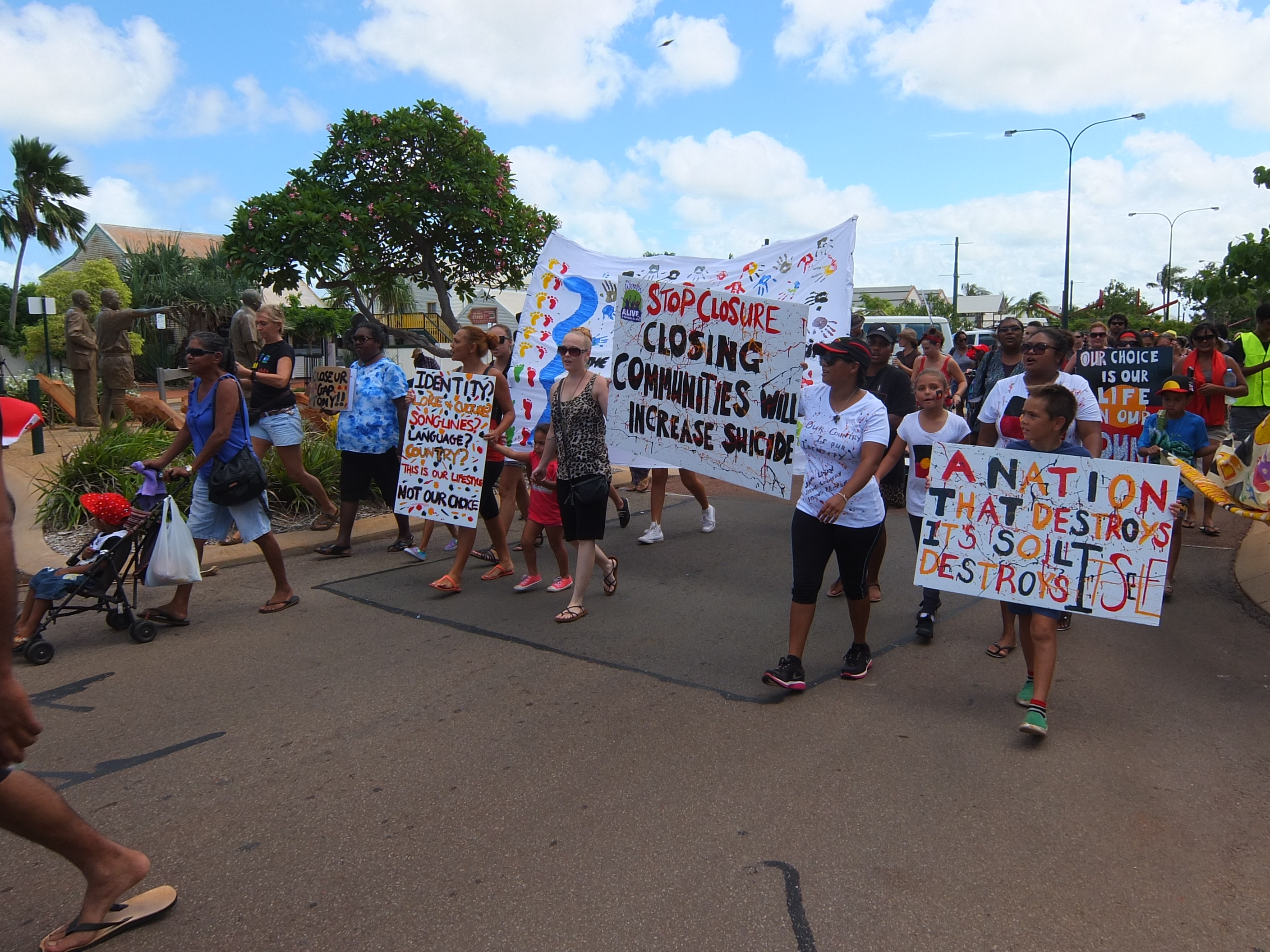 A view from the front showing dozens of people marching down the street in Broome. They hold large signs with messages protesting community closures.